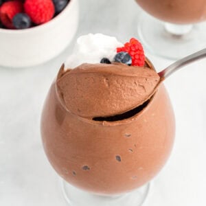 A glass filled with vegan chocolate mousse topped with whipped coconut cream and fresh berries, with a spoon scooping into the mousse and a bowl of mixed berries in the background.