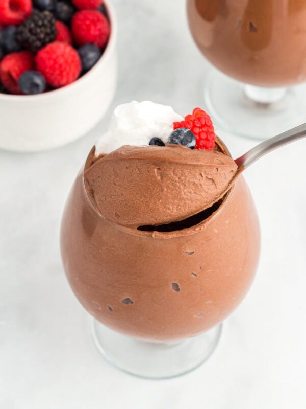 A glass filled with vegan chocolate mousse topped with whipped coconut cream and fresh berries, with a spoon scooping into the mousse and a bowl of mixed berries in the background.
