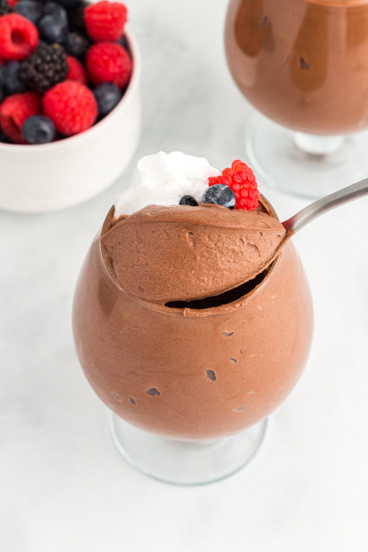 A glass filled with vegan chocolate mousse topped with whipped coconut cream and fresh berries, with a spoon scooping into the mousse and a bowl of mixed berries in the background.