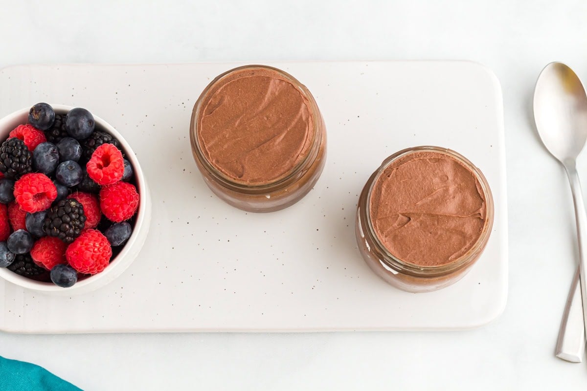 Two small jars filled with vegan chocolate mousse set on a white tray next to a bowl of fresh raspberries, blueberries, and blackberries.
