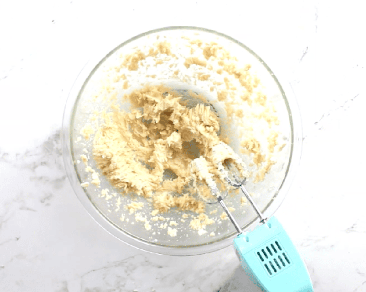 Creamed butter and sugars in a glass mixing bowl with an electric hand mixer on a marble countertop.
