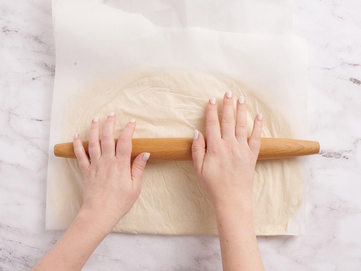 Hands rolling out the gluten free dough between two sheets of parchment paper.