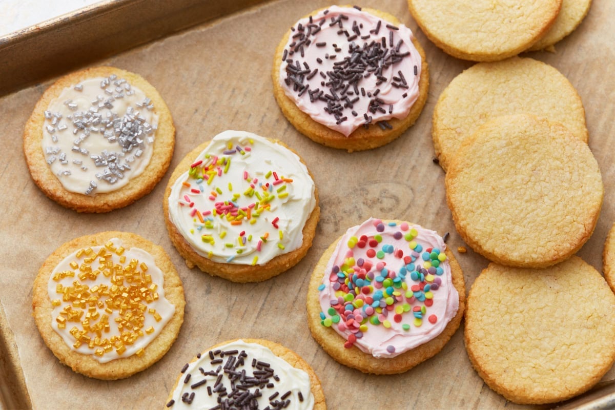 Almond flour sugar cookies decorated with colorful frosting and sprinkles next to plain baked cookies on a parchment-lined sheet.