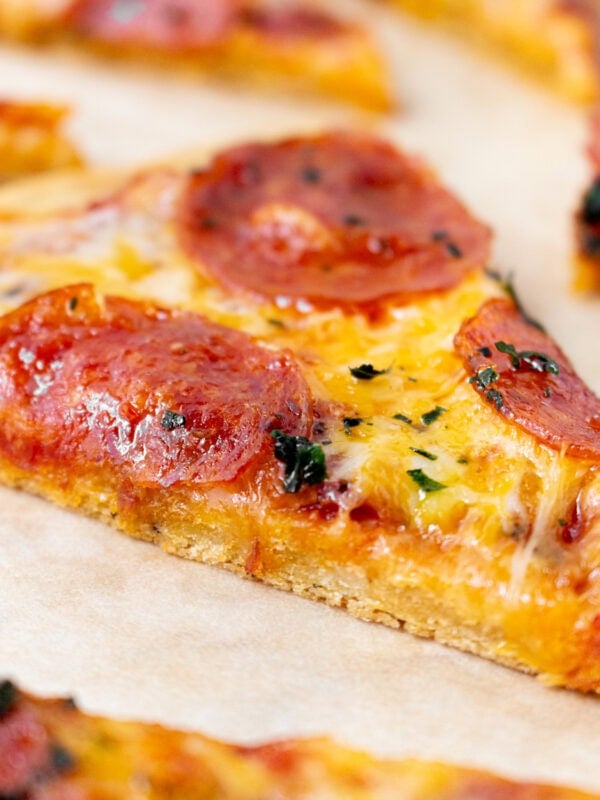 Slice of chickpea pizza crust topped with melted cheese and pepperoni on parchment paper