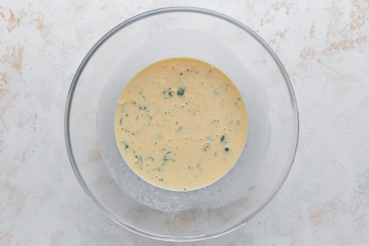 Whisked egg mixture with herbs in a glass bowl before baking a crustless ham and cheese quiche.