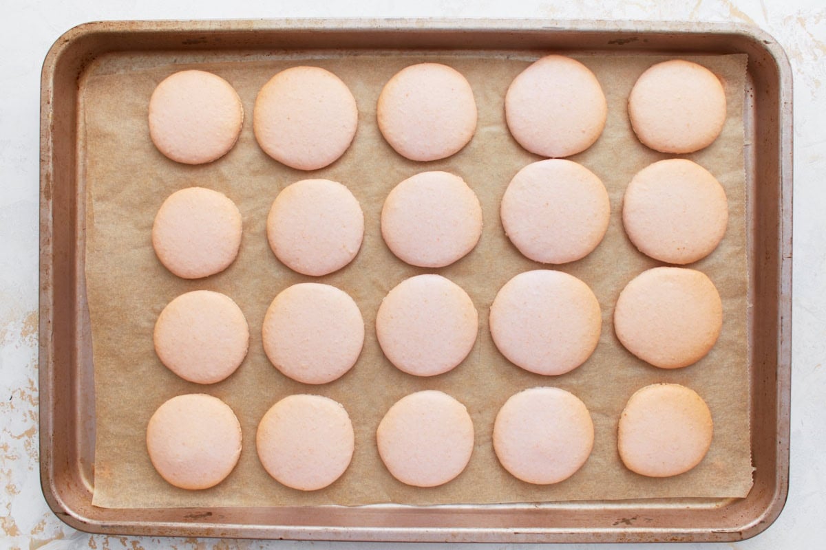 Baked pink macaron shells cooling on a parchment-lined tray, smooth and evenly shaped.