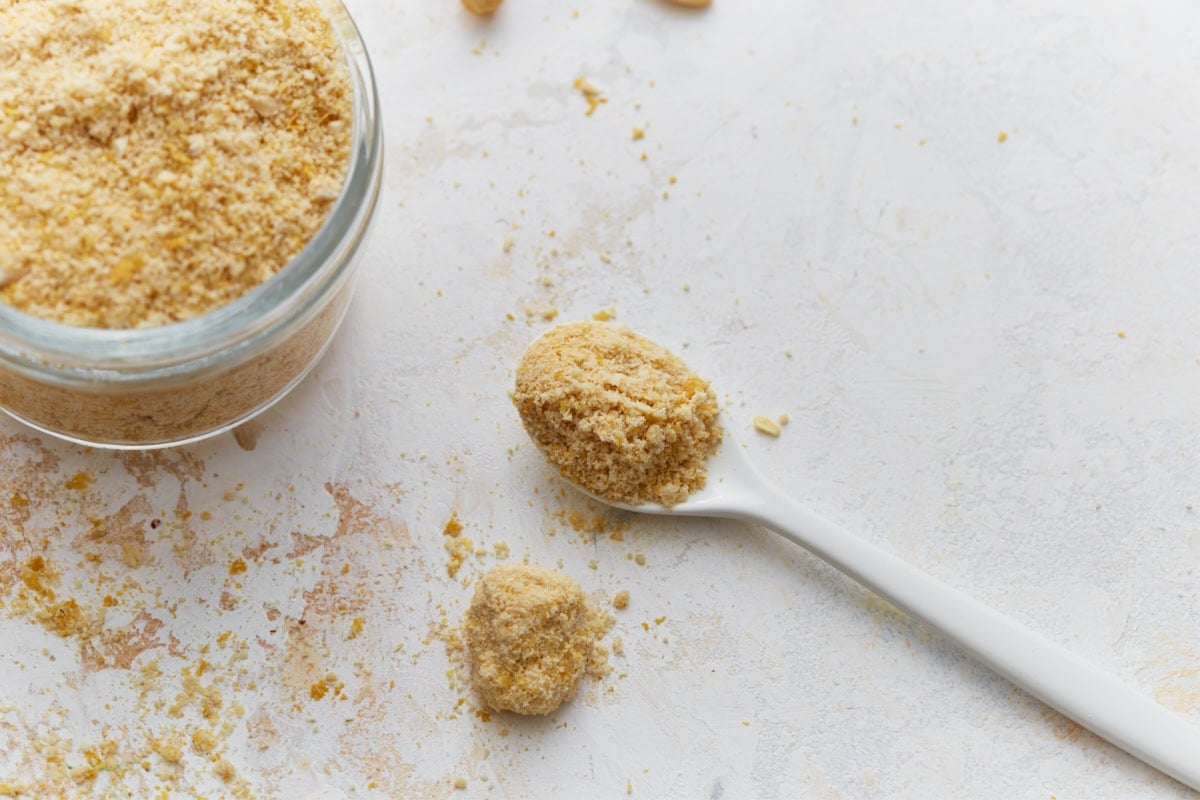 Spoonful of vegan parmesan cheese next to an open jar on a light background.
