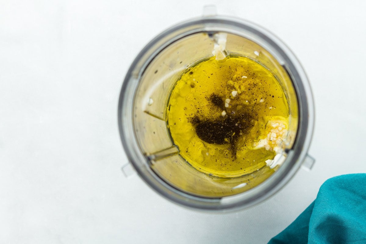 Ingredients for dairy-free Caesar dressing in a blender including olive oil, garlic, lemon juice, Dijon mustard, and black pepper