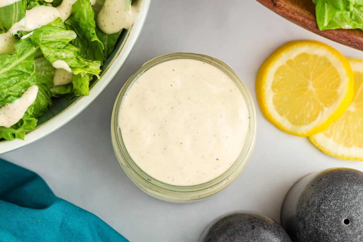 Small glass jar filled with creamy dairy-free Caesar dressing next to fresh romaine lettuce and lemon slices