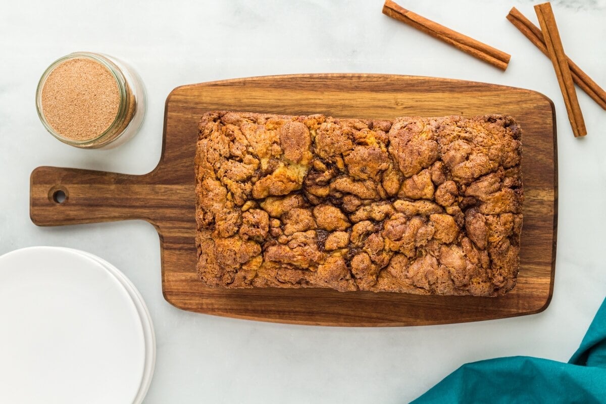 Whole loaf of gluten-free cinnamon bread on a wooden cutting board with cinnamon sugar and cinnamon sticks.