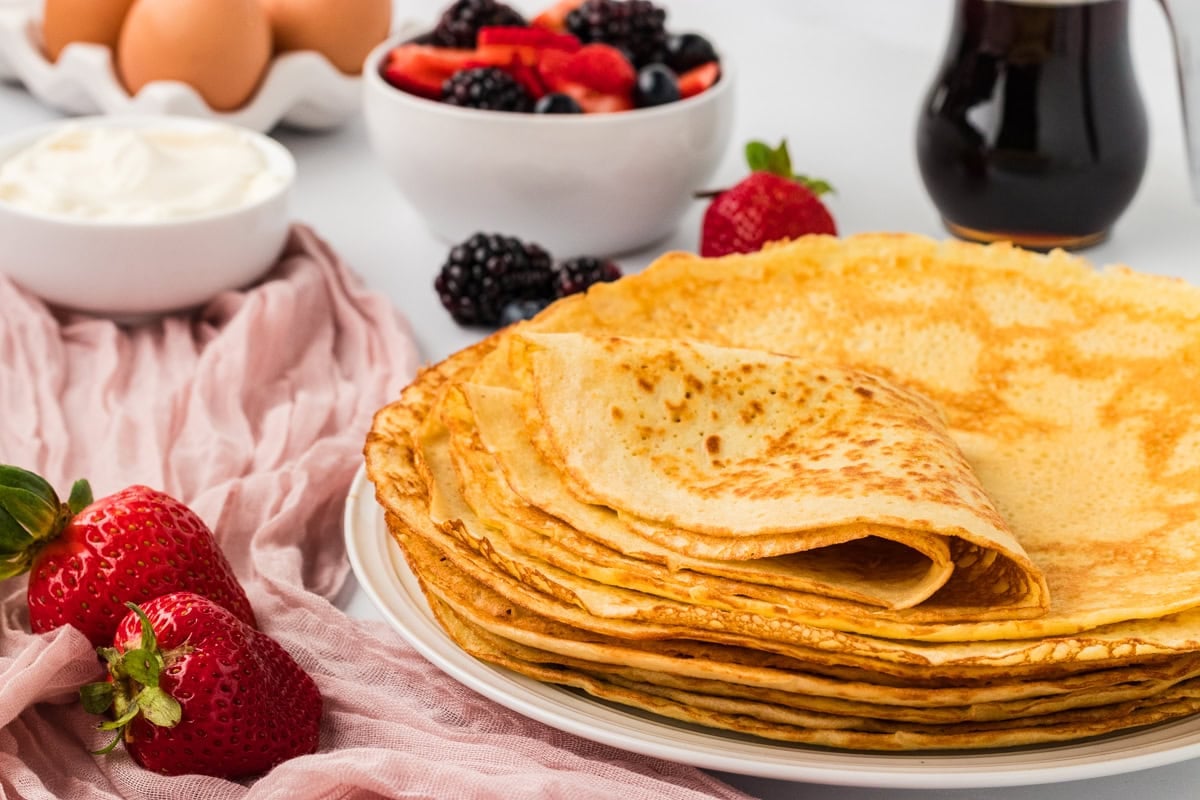 Stack of folded almond flour crepes with strawberries, blackberries, and maple syrup nearby