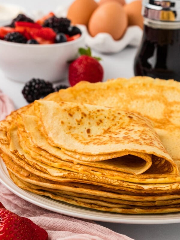 Stack of almond flour crepes on a white plate with fresh berries, eggs, and maple syrup in the background