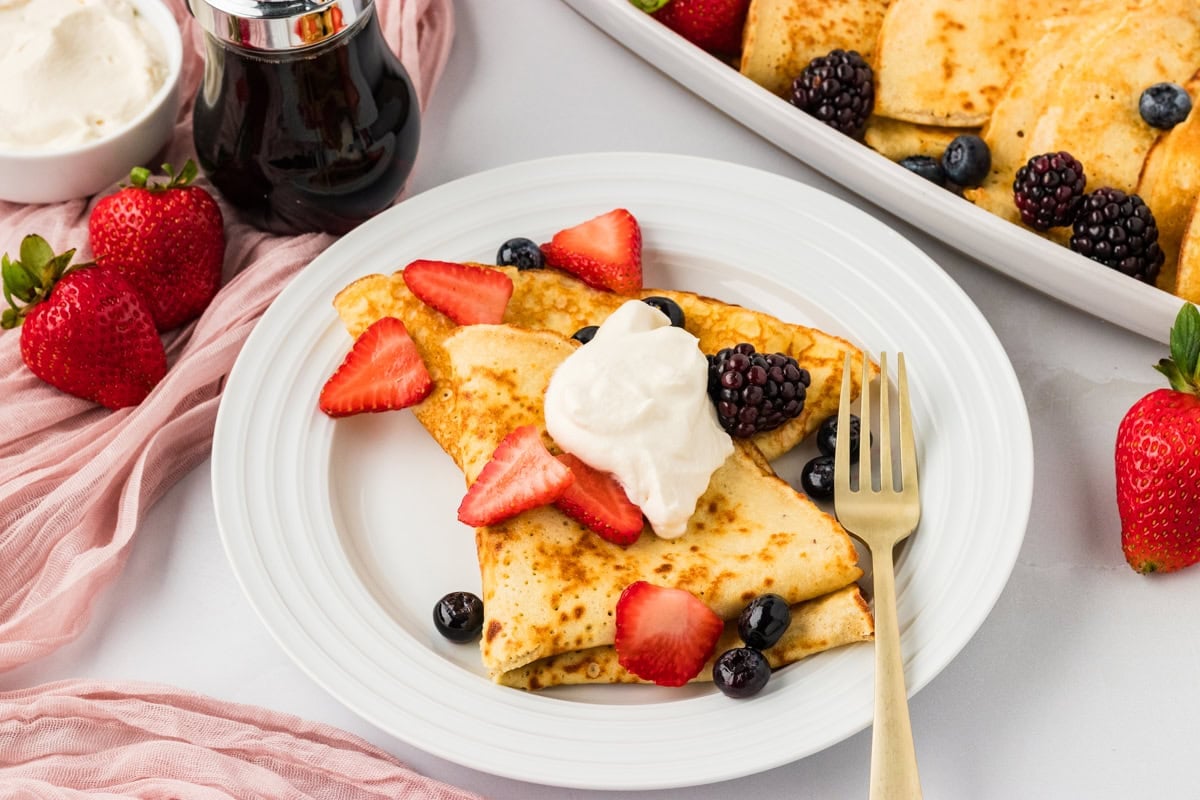 Stack of folded almond flour crepes with strawberries, blackberries, and maple syrup nearby