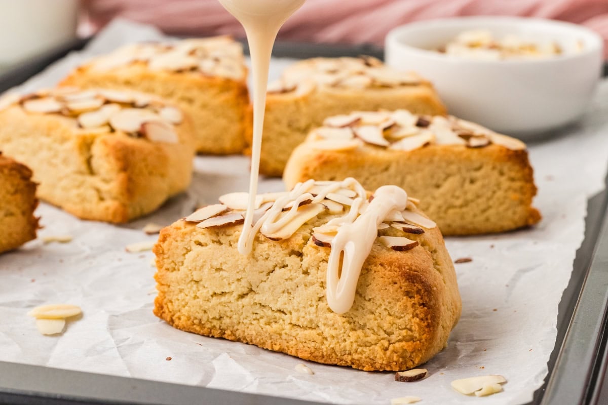 Vanilla icing being drizzled over warm almond flour scone on baking tray