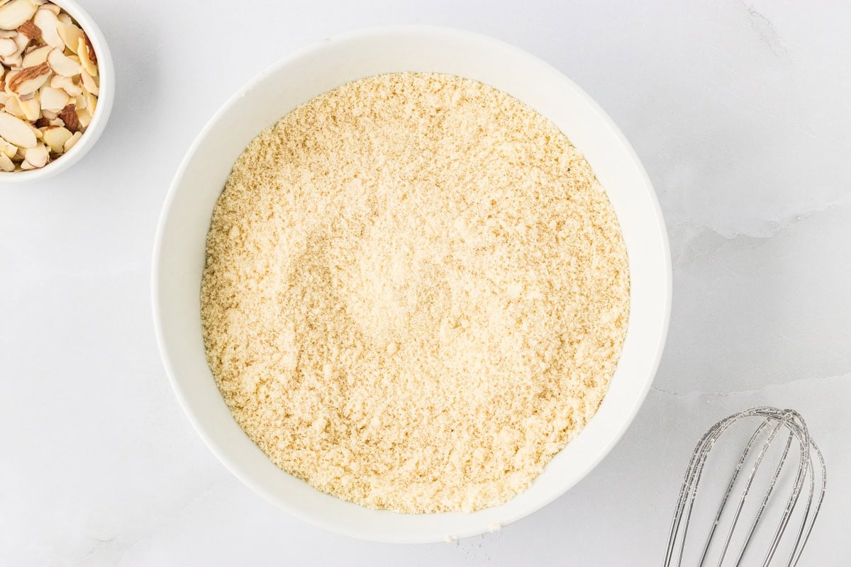 Bowl of fine almond flour with whisk and sliced almonds on white surface