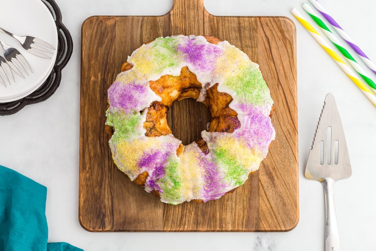 Gluten free king cake topped with vanilla glaze and purple, yellow, and green sanding sugars on a wooden board.