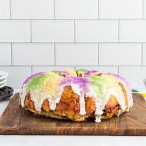 Gluten free king cake topped with white vanilla glaze and purple, green, and gold sanding sugar on a wooden board.