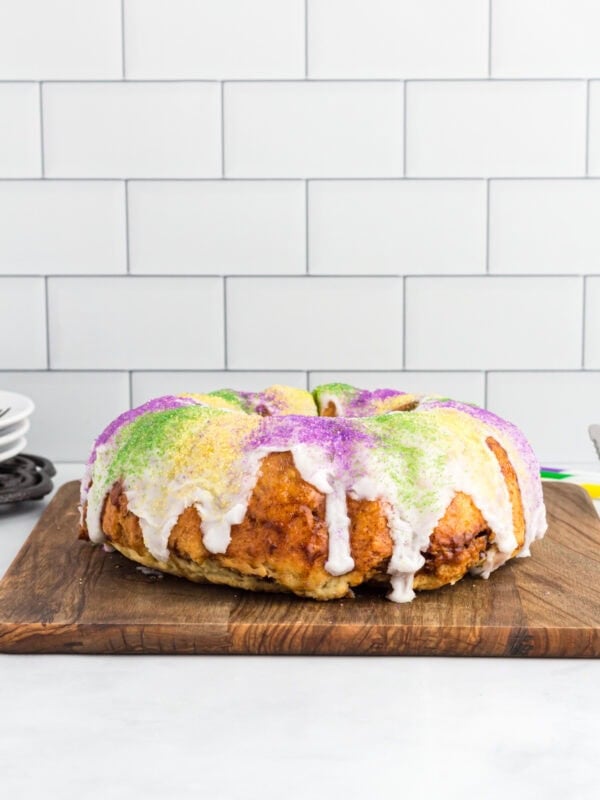 Gluten free king cake topped with white vanilla glaze and purple, green, and gold sanding sugar on a wooden board.