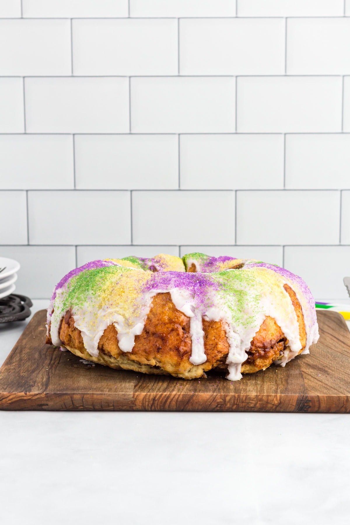 Gluten free king cake topped with white vanilla glaze and purple, green, and gold sanding sugar on a wooden board.