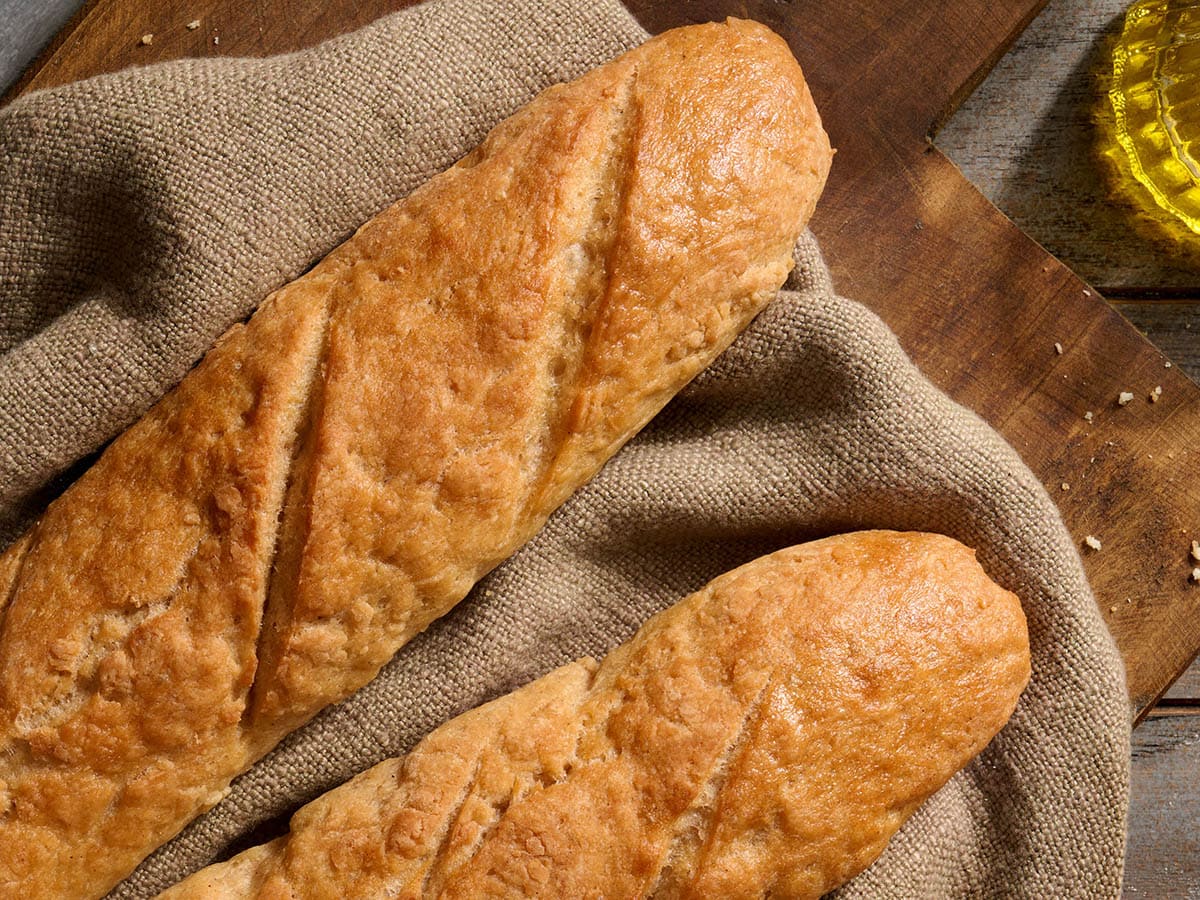 Two homemade gluten-free baguettes with golden crust resting on a linen cloth on a wooden board.
