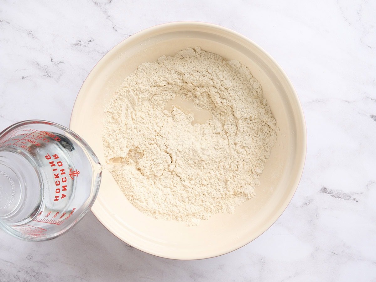 Warm water being poured into gluten-free flour mixture in a mixing bowl to form baguette dough.