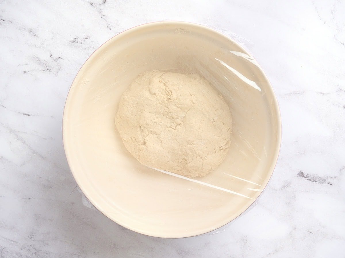 Gluten-free baguette dough covered tightly with plastic wrap in a bowl before the first rise.
