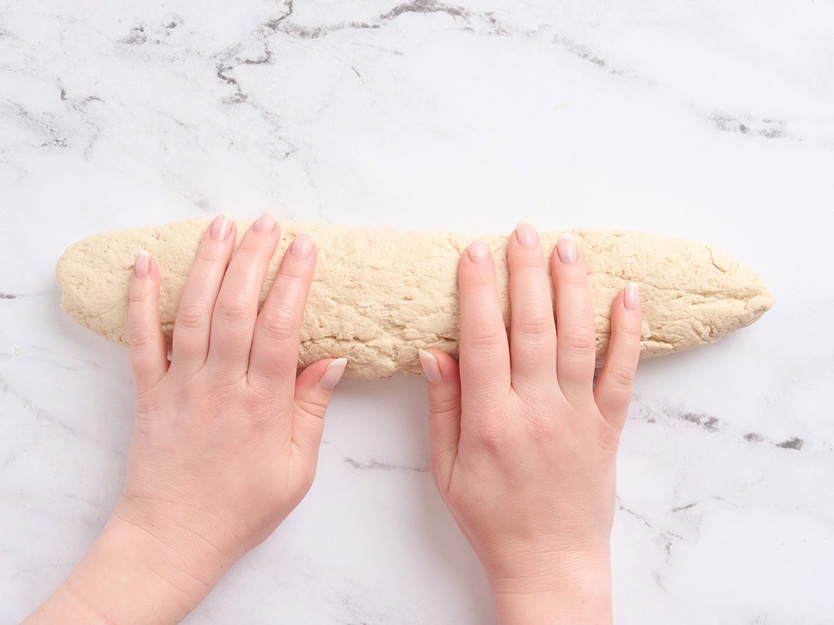 Hands gently shaping gluten-free dough into a long 12-inch baguette log on a lightly floured surface.