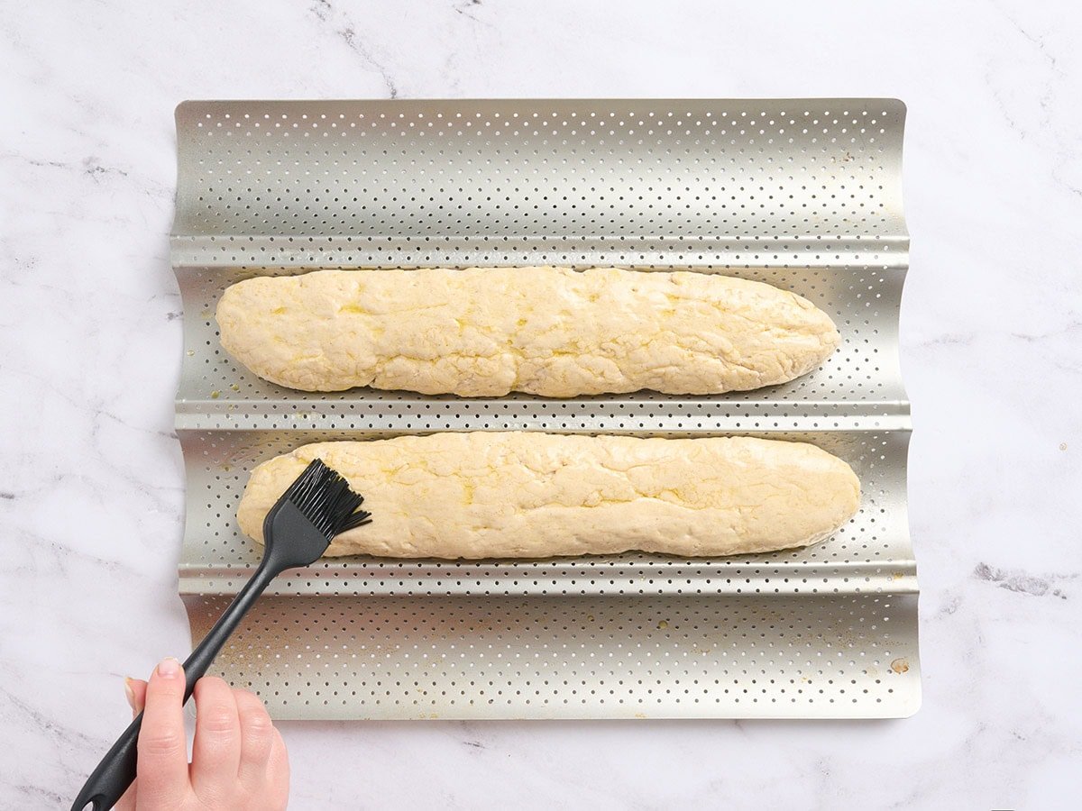 Sharp knife scoring diagonal slashes into gluten-free baguette dough before baking.