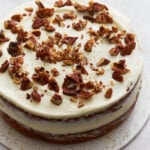 Almond flour carrot cake layered and frosted with cream cheese frosting, topped with chopped pecans on a white cake stand.