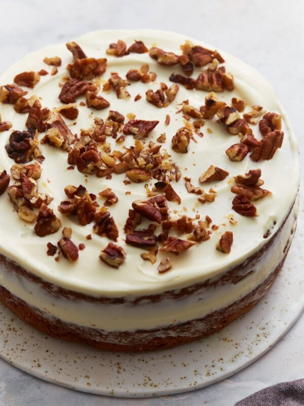 Almond flour carrot cake layered and frosted with cream cheese frosting, topped with chopped pecans on a white cake stand.