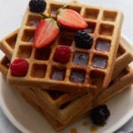 Stacked buckwheat waffles topped with strawberries, raspberries, blackberries, and maple syrup on a white plate