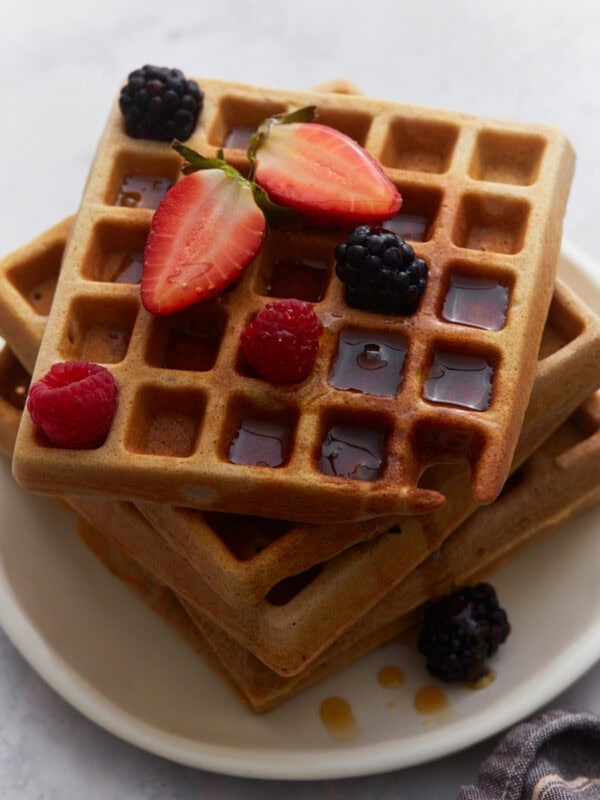 Stacked buckwheat waffles topped with strawberries, raspberries, blackberries, and maple syrup on a white plate