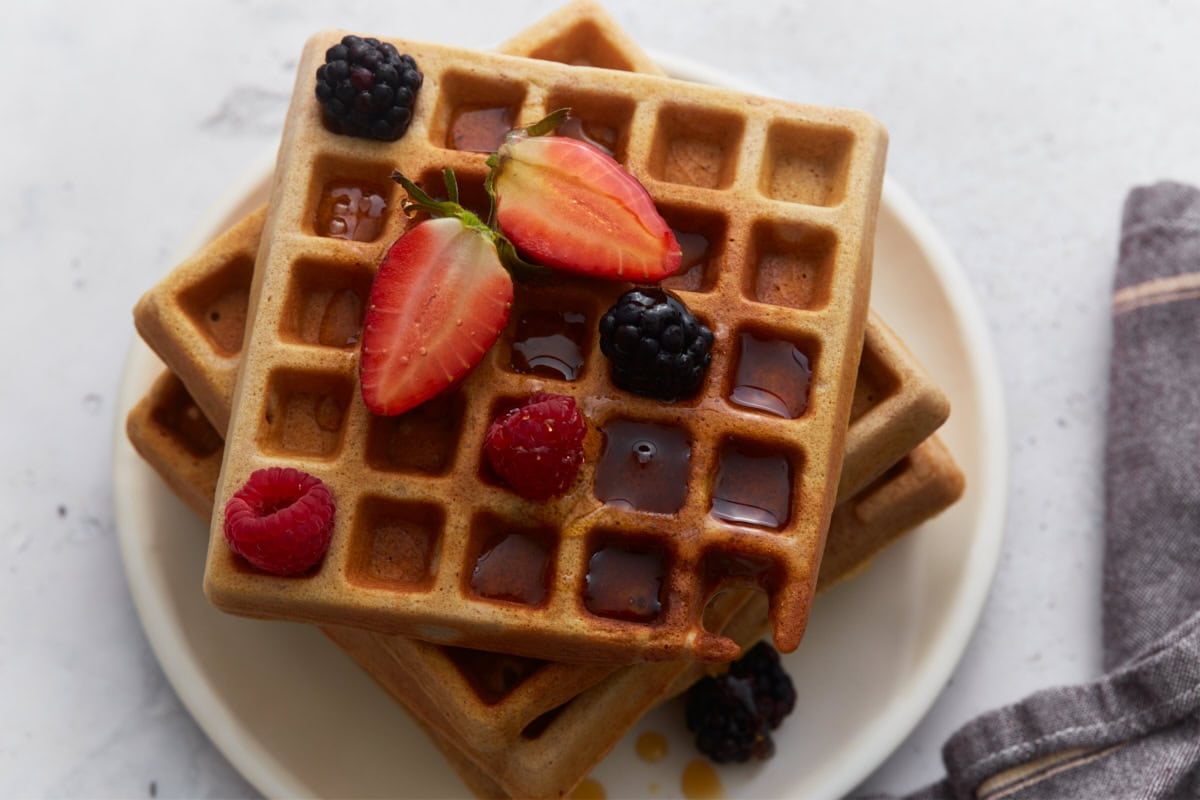Buckwheat waffles stacked on a plate with fresh berries and maple syrup drizzle