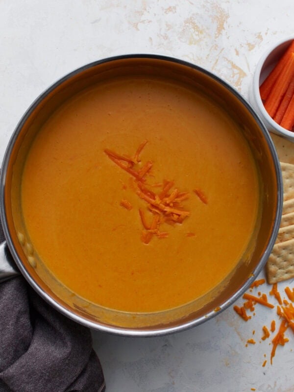 Gluten-free beer cheese dip finished in saucepan with shredded cheddar on top, served with crackers and carrots