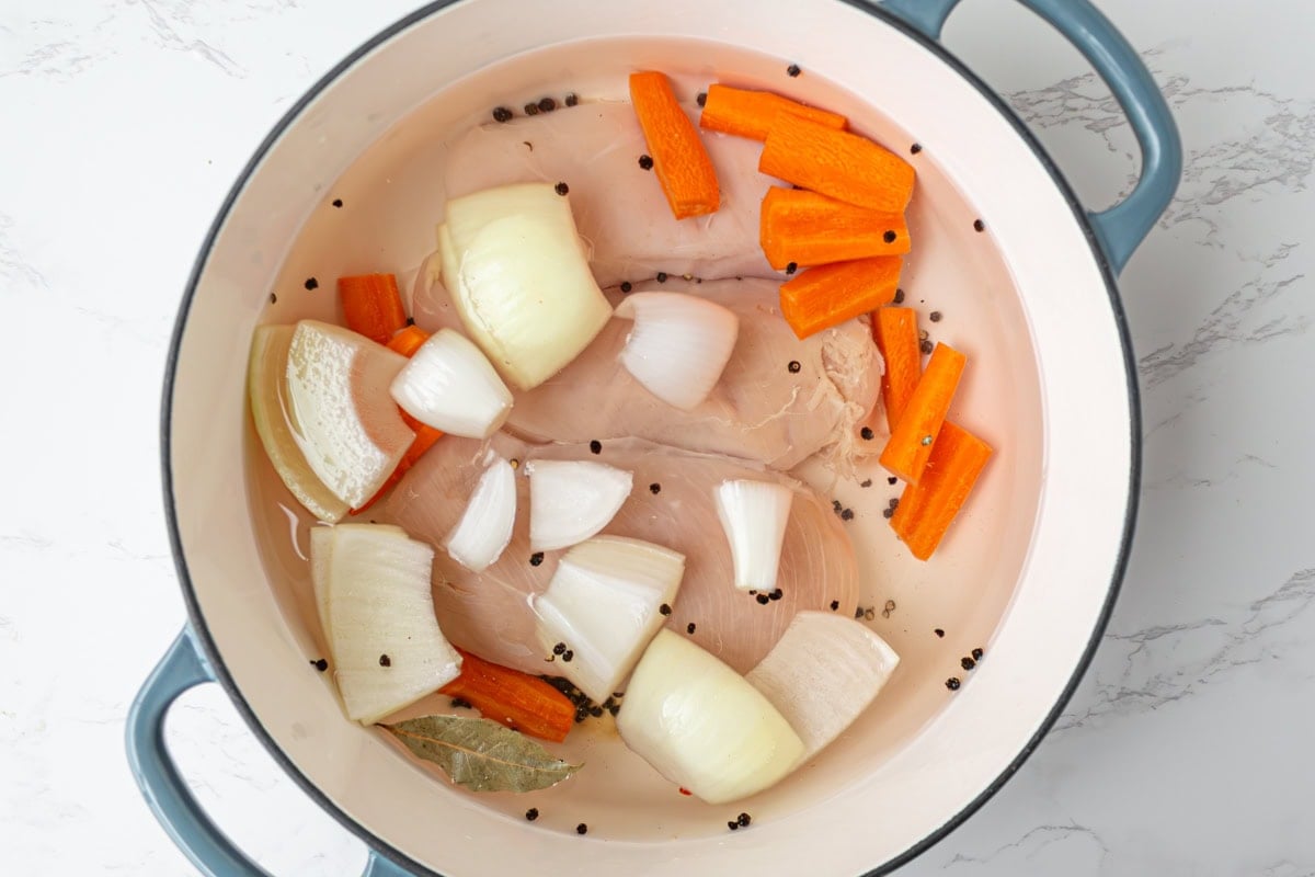 Chicken simmering in a Dutch oven with carrot chunks, onion quarters, bay leaf, and peppercorns to make homemade broth.