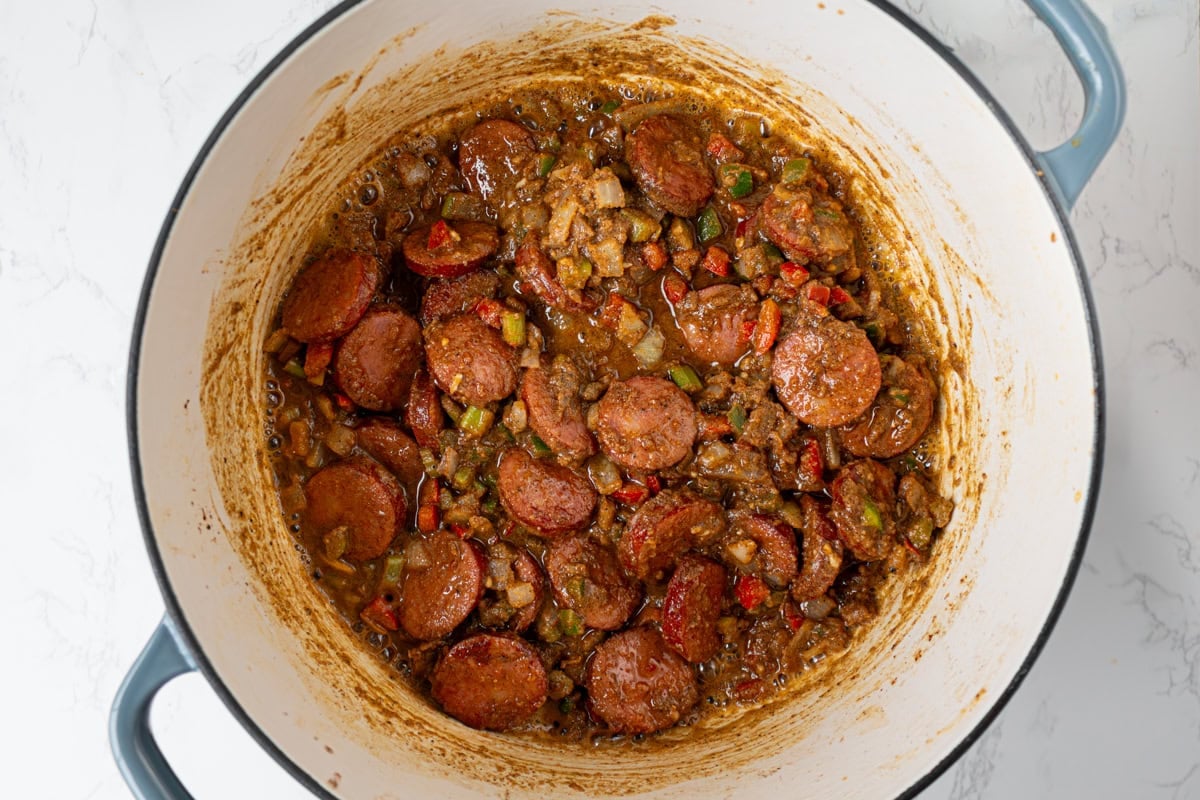 Andouille sausage and sautéed vegetables coated in dark roux inside a Dutch oven as the gumbo base forms.
