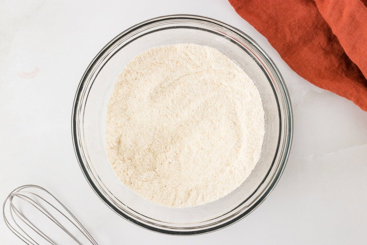 Mixed dry ingredients for gluten free hush puppies in a glass bowl with a whisk nearby.
