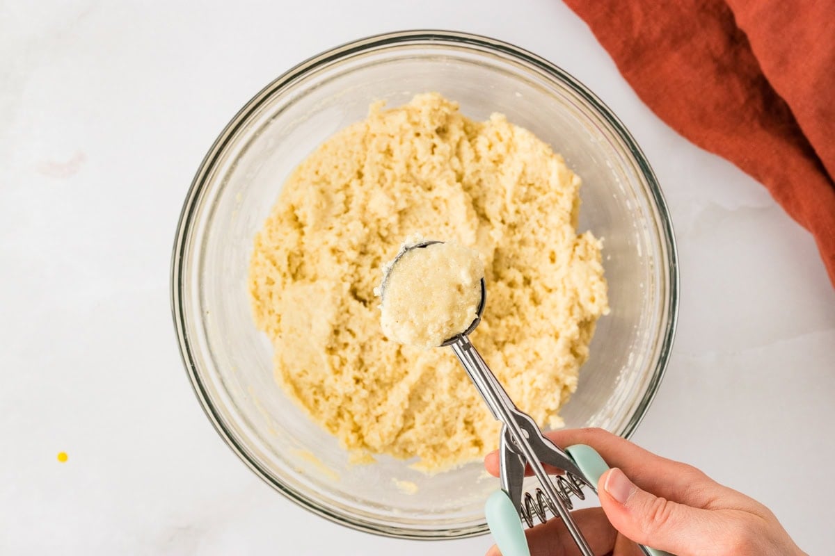 Cookie scoop portioning gluten free hush puppy batter from a mixing bowl.