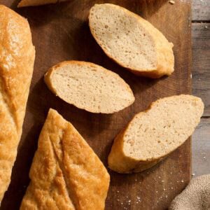 Sliced gluten-free baguette with golden crust and soft crumb on a wooden board next to olive oil for dipping.