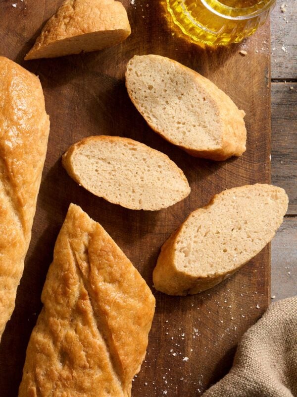 Sliced gluten-free baguette with golden crust and soft crumb on a wooden board next to olive oil for dipping.