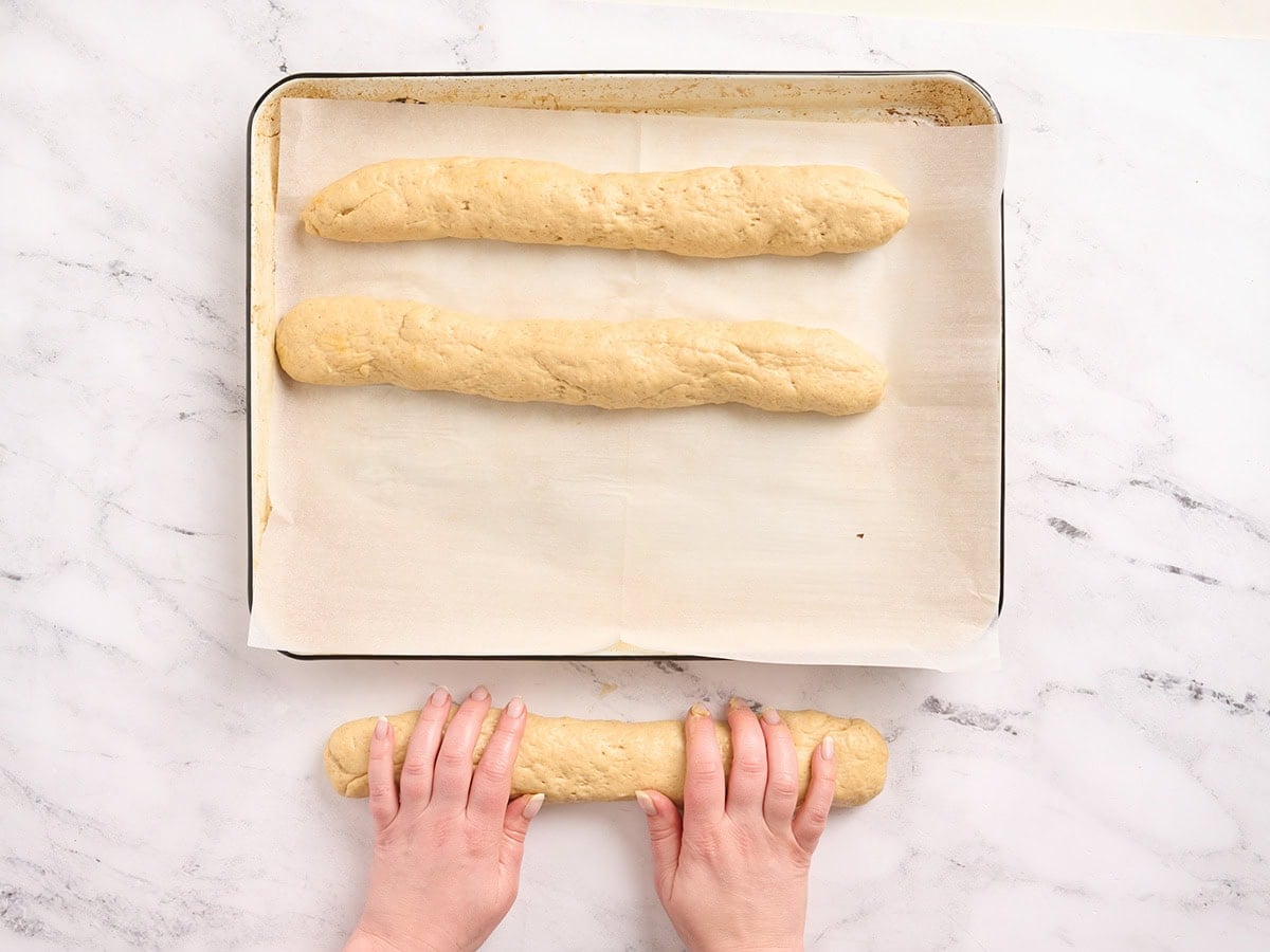Hands rolling gluten free challah bread dough into three ropes on a parchment lined baking sheet for braiding.