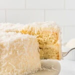 Gluten-free coconut cake slice being lifted from a frosted coconut layer cake covered in shredded coconut.