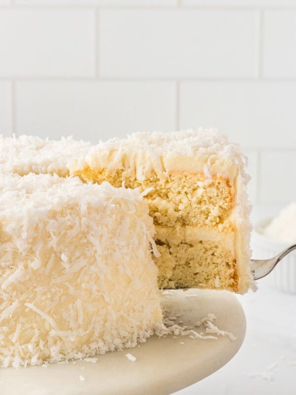Gluten-free coconut cake slice being lifted from a frosted coconut layer cake covered in shredded coconut.