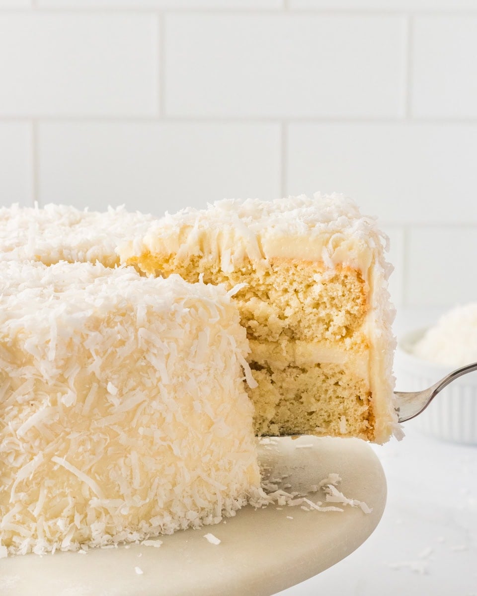 Gluten-free coconut cake slice being lifted from a frosted coconut layer cake covered in shredded coconut.