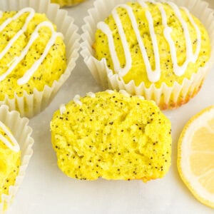 Gluten-free lemon poppy seed muffins with lemon glaze in white liners, one muffin sliced open to show moist crumb and poppy seeds