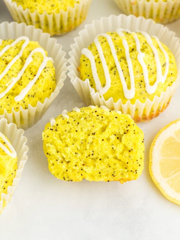Gluten-free lemon poppy seed muffins with lemon glaze in white liners, one muffin sliced open to show moist crumb and poppy seeds
