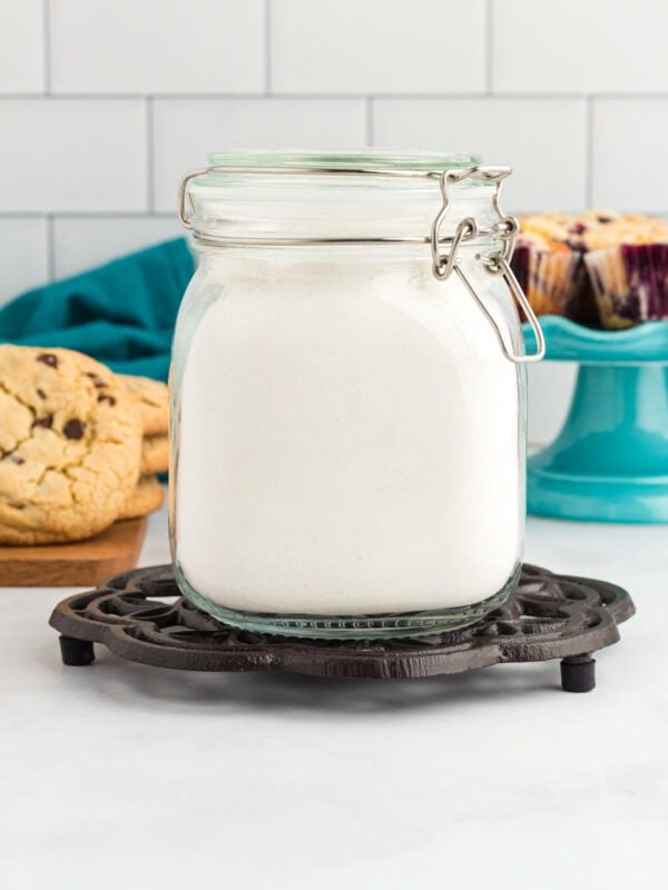 Homemade gluten free flour blend stored in an airtight glass jar with cookies and muffins in the background