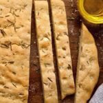 Sliced gluten-free focaccia bread with rosemary and flaky salt served on a wooden board with olive oil for dipping