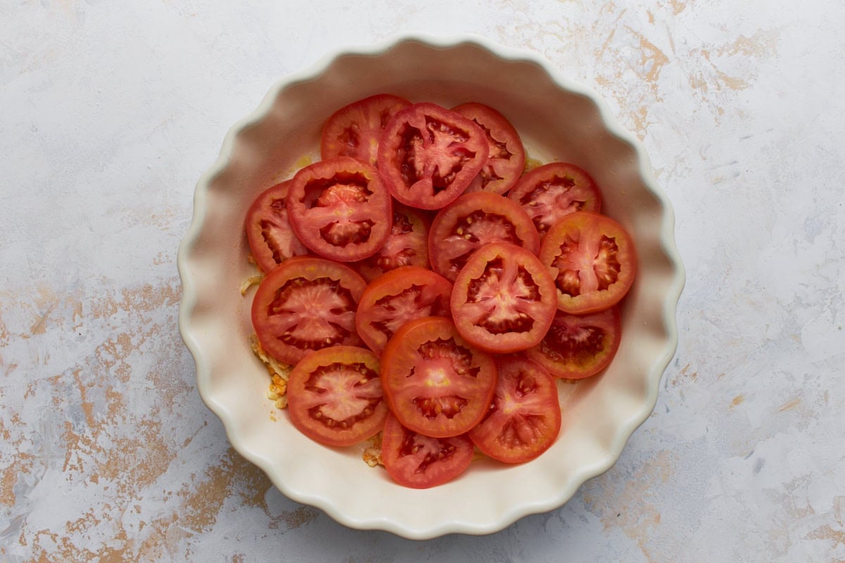 Layer of sliced tomatoes arranged over the cheese mixture in a white pie dish before baking.