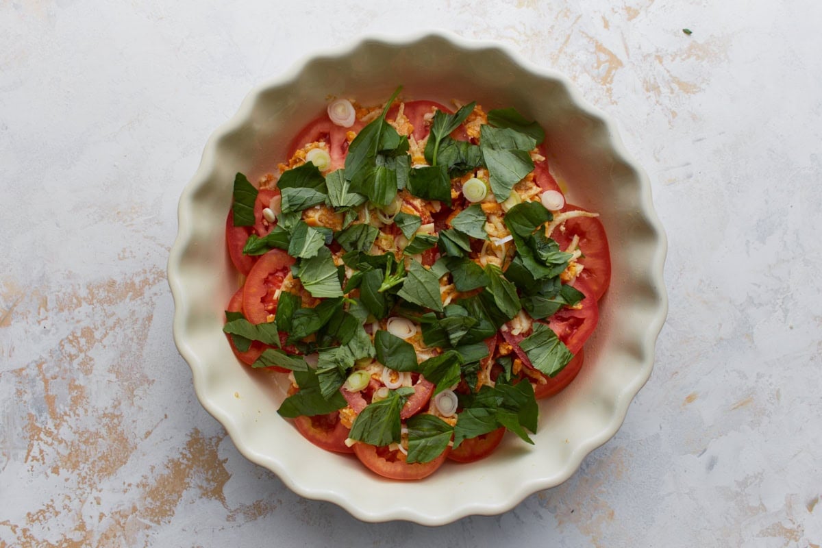 Overhead view of crustless tomato pie before baking with layers of sliced tomatoes, cheese mixture, fresh basil, and green onions in a white pie dish.